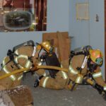 Two firefighters crawling on the floor of the training house holding a firehose
