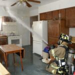 A firefighter spraying water from a firehose onto the ceiling above the stove in the training house kitchen.