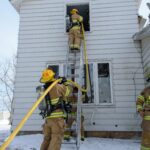 Firefighters at a training house. Two are holding a firehose. The first is up on a ladder to the second floor of the house while the other at the base of the ladder.