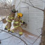 Two firefighters cutting a square opening with a saw into the side of the training house.