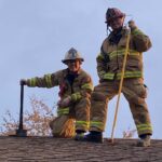 Two firefighters standing on a roof. One is holding a pole hook and the other holding an axe.